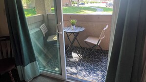 Un balcón con puerta de cristal, una mesita redonda pequeña, dos sillas de mimbre y una planta en maceta.