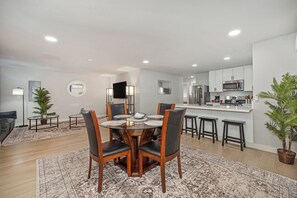 A modern kitchen with stainless steel appliances, a dining area with a glass table and leather chairs, and a living room with a sofa and a floor lamp.