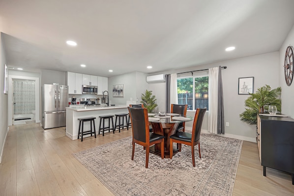 Una cocina moderna con armarios blancos, una isla central y una zona de comedor con mesa de vidrio y sillas de madera.