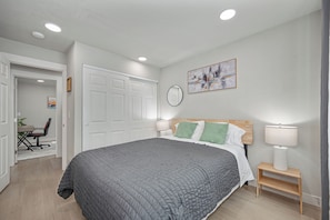 Quarto com uma cama grande, escrivaninha com cadeira, porta e quadro na parede.