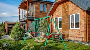 Una casa de madera de dos pisos con un balcón, un conjunto de columpios y un jardín bien cuidado.