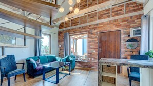 Amplia sala de estar con pared de ladrillo, vigas de madera y un ventanal grande. Hay un sofá, una mesa de centro y una mesa de comedor con sillas.