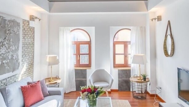 A spacious living room with a high ceiling, wooden beams, a white sofa, a white armchair, a coffee table with flowers, and a television. - San Bernardo ático de 1 dormitorio (Sevilla)