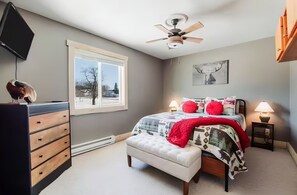A bedroom with a bed, bedside tables, a dresser, a ceiling fan, a window, and a wall-mounted TV.