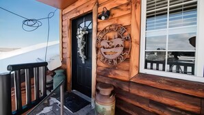A wooden cabin with a black door, a welcome sign, a window with blinds, and a metal container on the porch. - Boyne Mtn Views! 2BR w/ Patio & Fire Pit Access (Boyne Falls)