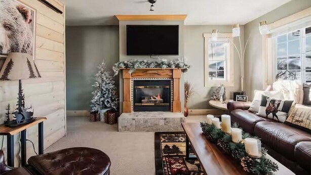 A cozy living room with a fireplace, a leather sofa, and a television mounted on the wall. - Boyne Mtn Views! 2BR w/ Patio & Fire Pit Access (Boyne Falls)