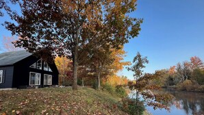 Eine Hütte mit großem Fenster und Tür, umgeben von Herbstlaub und einem stillen See.