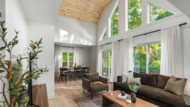 Ein geräumiges Wohnzimmer mit hohen Decken, großen Fenstern, einem gemütlichen Sofa, einem Couchtisch und einem Essbereich im Hintergrund.