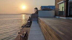 A wooden deck with chairs overlooking a calm sea, a wooden building with large windows, and a distant bridge.