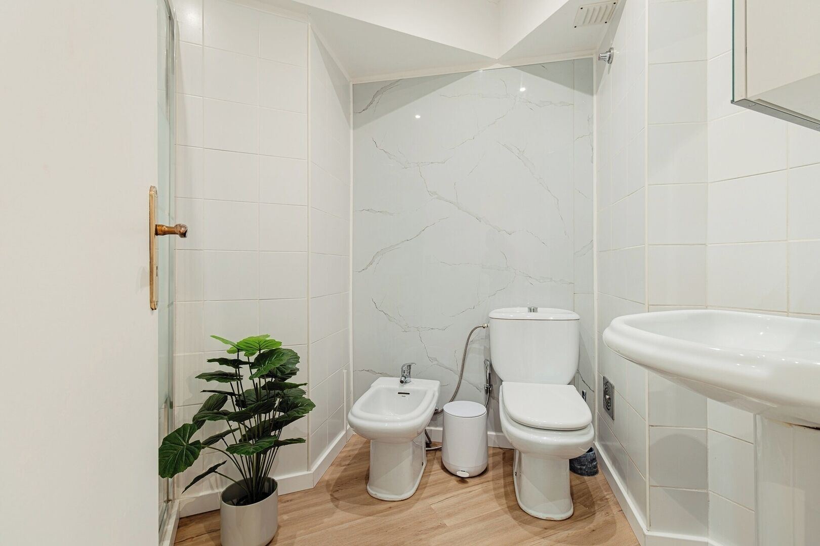 Een moderne badkamer met witte marmeren wanden, een bidet, toilet, wastafel en een potplant.