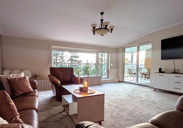 A spacious living room with a brown leather sofa, a wooden coffee table, and a chandelier. Large windows provide natural light and a view of the outdoors. - Comfortable Apartment, Sauna Included in the Price (Kołobrzeg)