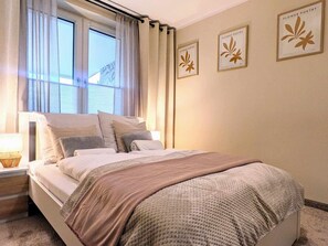 A bedroom with a bed, a nightstand, a window with curtains, and three framed pictures on the wall. - Comfortable Apartment, Sauna Included in the Price (Kołobrzeg)