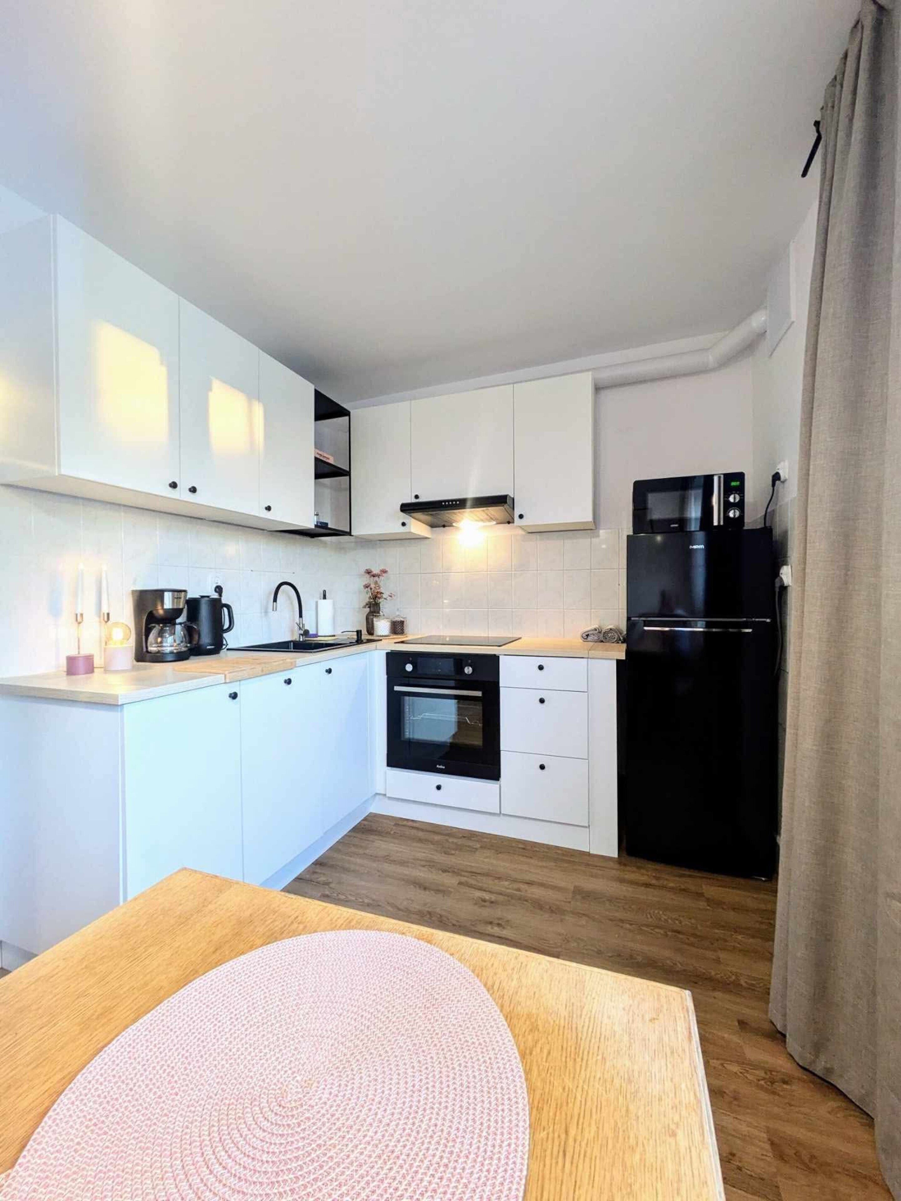 A modern kitchen with white cabinets, a black oven, a black refrigerator, and a wooden dining table with a pink placemat.