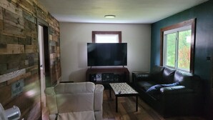 Una sala de estar con una televisión de pantalla plana, un sofá de cuero, un sillón blanco y una mesa de centro de madera.