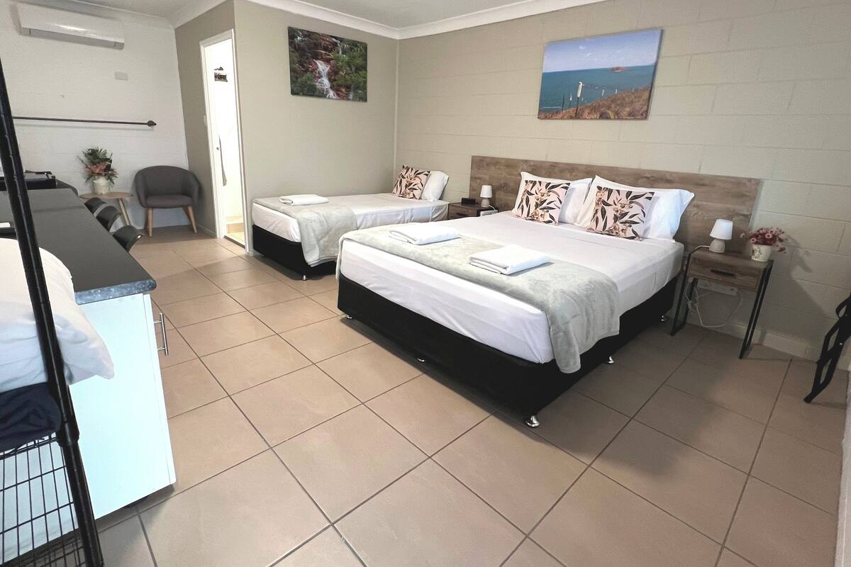 A hotel room with two beds, a desk, a chair, a small table with a lamp, and two paintings on the wall.