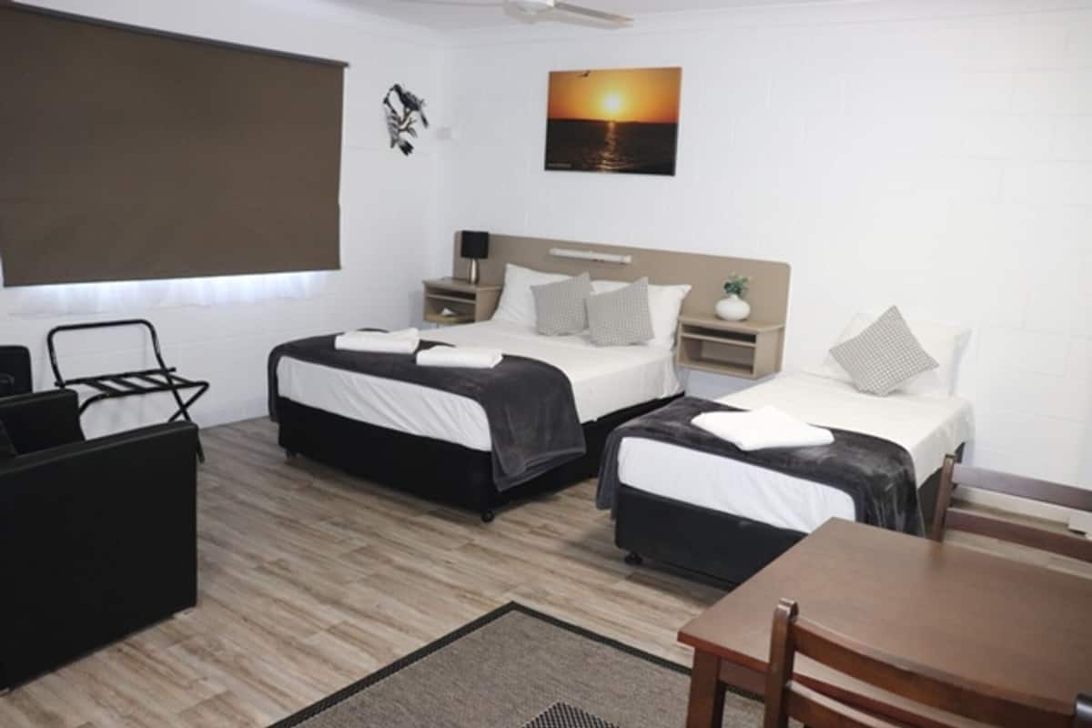 A hotel room with two beds, a desk, a chair, a small table, a ceiling fan, a wall-mounted TV, and a painting of a sunset.
