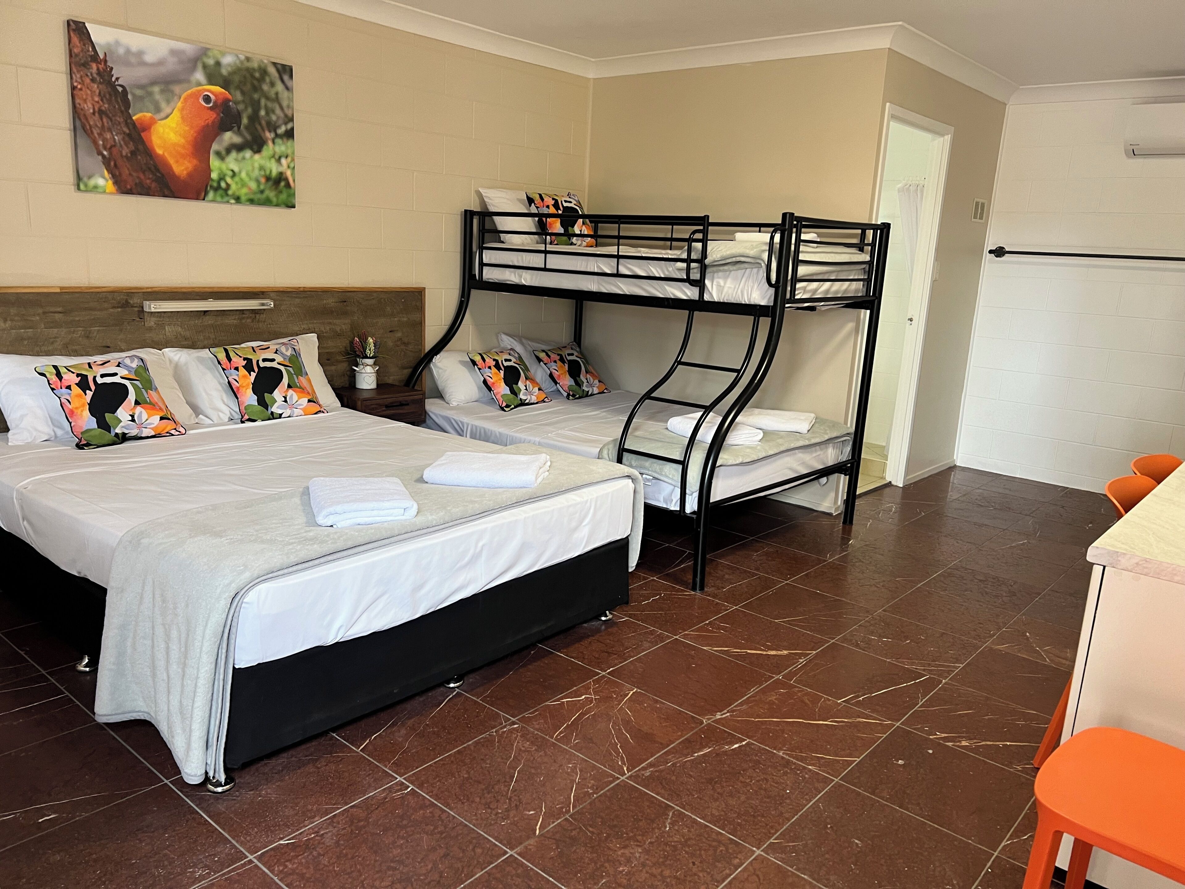 A hotel room with two beds, one with a headboard and one with a bunk bed. There is a small table with a chair, a potted plant, and a painting on the wall.