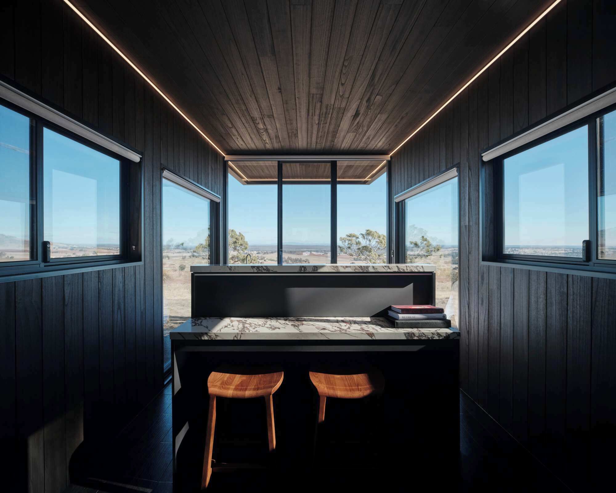 A modern interior with a desk, two wooden chairs, and large windows offering a view of the landscape.