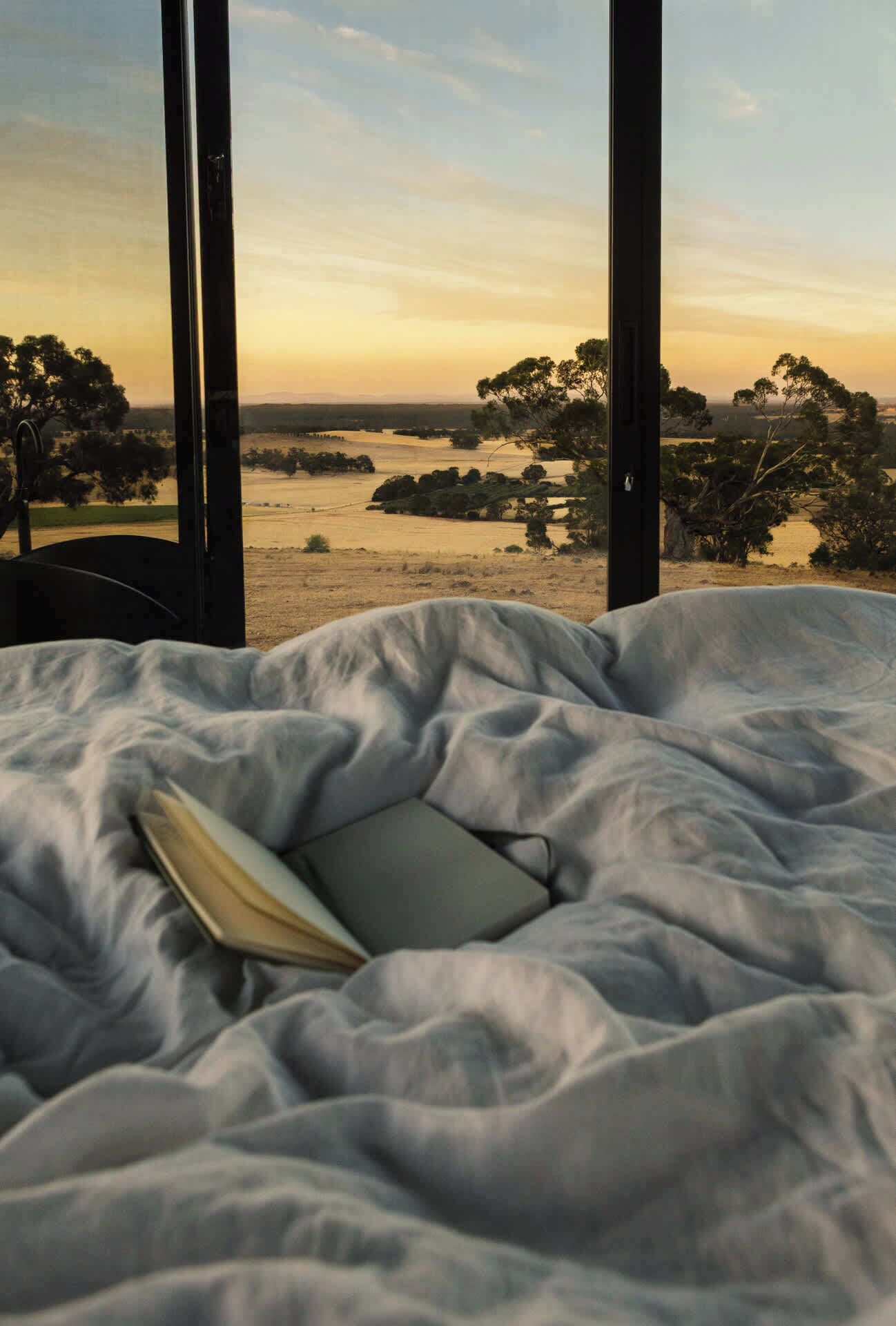 A bed with a book, large windows, and a view of trees and water.