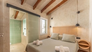 A bedroom with a bed, bedside tables, a window, and a bathroom with a shower. - Can Carnero | House in Menorca with sea views (Cap D'Artruix (Ciutadella De Menorca))