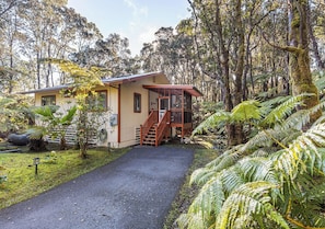 A house with a wooden deck, a gravel driveway, and surrounding lush greenery.
