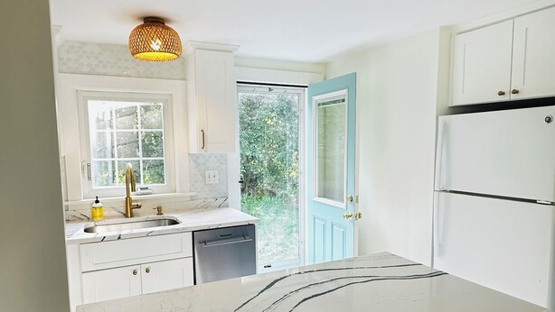 Een moderne keuken met witte kasten, een marmeren aanrechtblad en een ingebouwde vaatwasser. Er staan twee geweven krukken bij het eiland.
