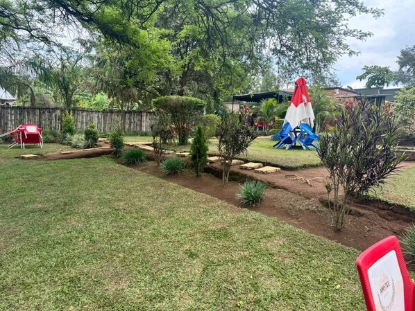 Un patio trasero con una carpa roja y blanca, un tobogán inflable azul y blanco, y una pequeña zona ajardinada con diversas plantas.