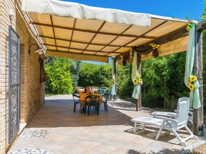 Eine überdachte Terrasse mit Tisch und Stühlen, umgeben von viel Grün.