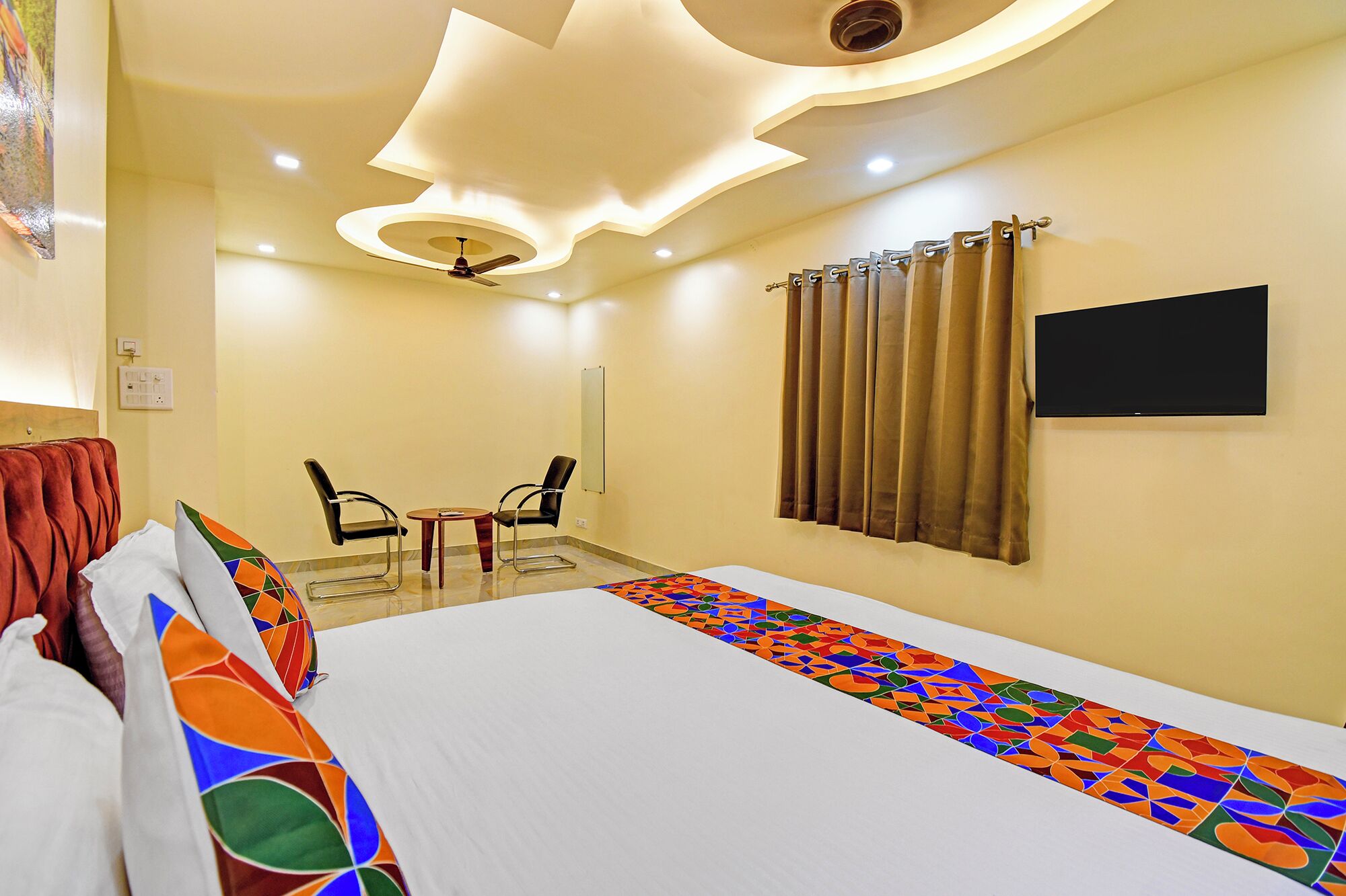 A hotel room with a bed, a TV, a ceiling fan, and a small table with chairs.
