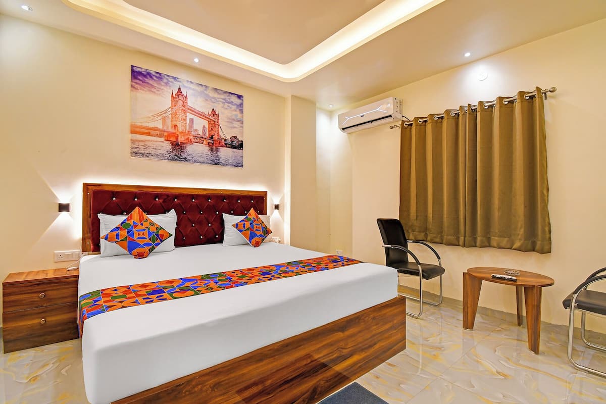 A hotel room with a large bed, a headboard, a painting of a bridge, an air conditioning unit, a chair, a small table, and a curtain.