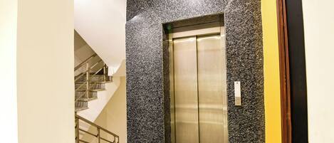 Un ascensor moderno con acabados metálicos, junto a una escalera con barandilla de metal.