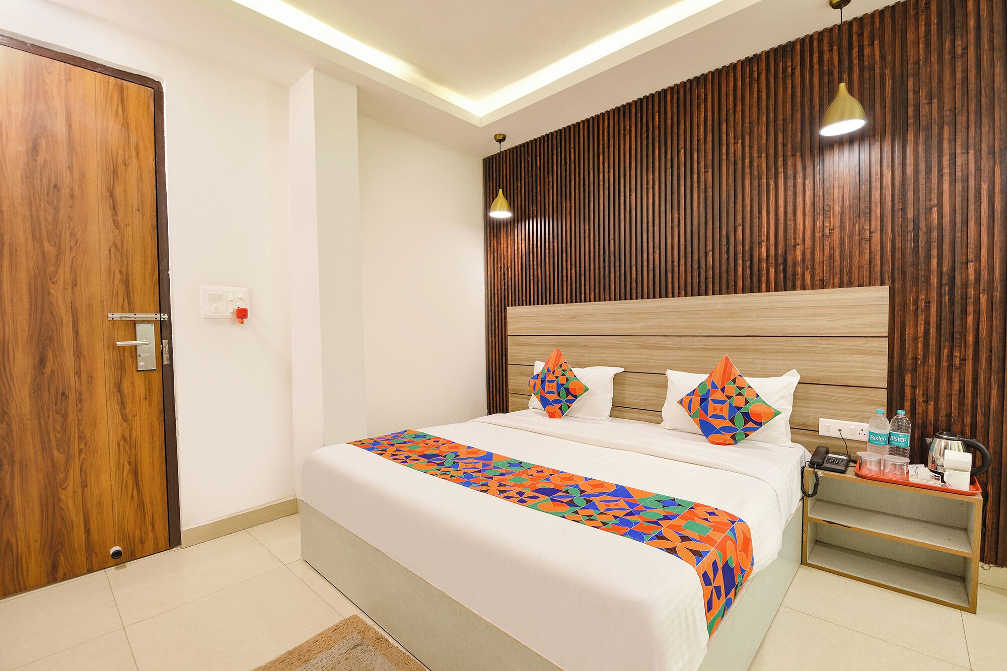 A hotel room with a wooden headboard, a bed with colorful geometric-patterned bedding, a bedside table with a phone and water bottles, and a wooden door.