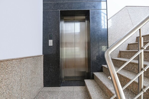 An elevator with a metallic interior, a control panel with a green indicator, and a staircase with metal railings. - FabHotel Ganeshwaram 45 (Noida)