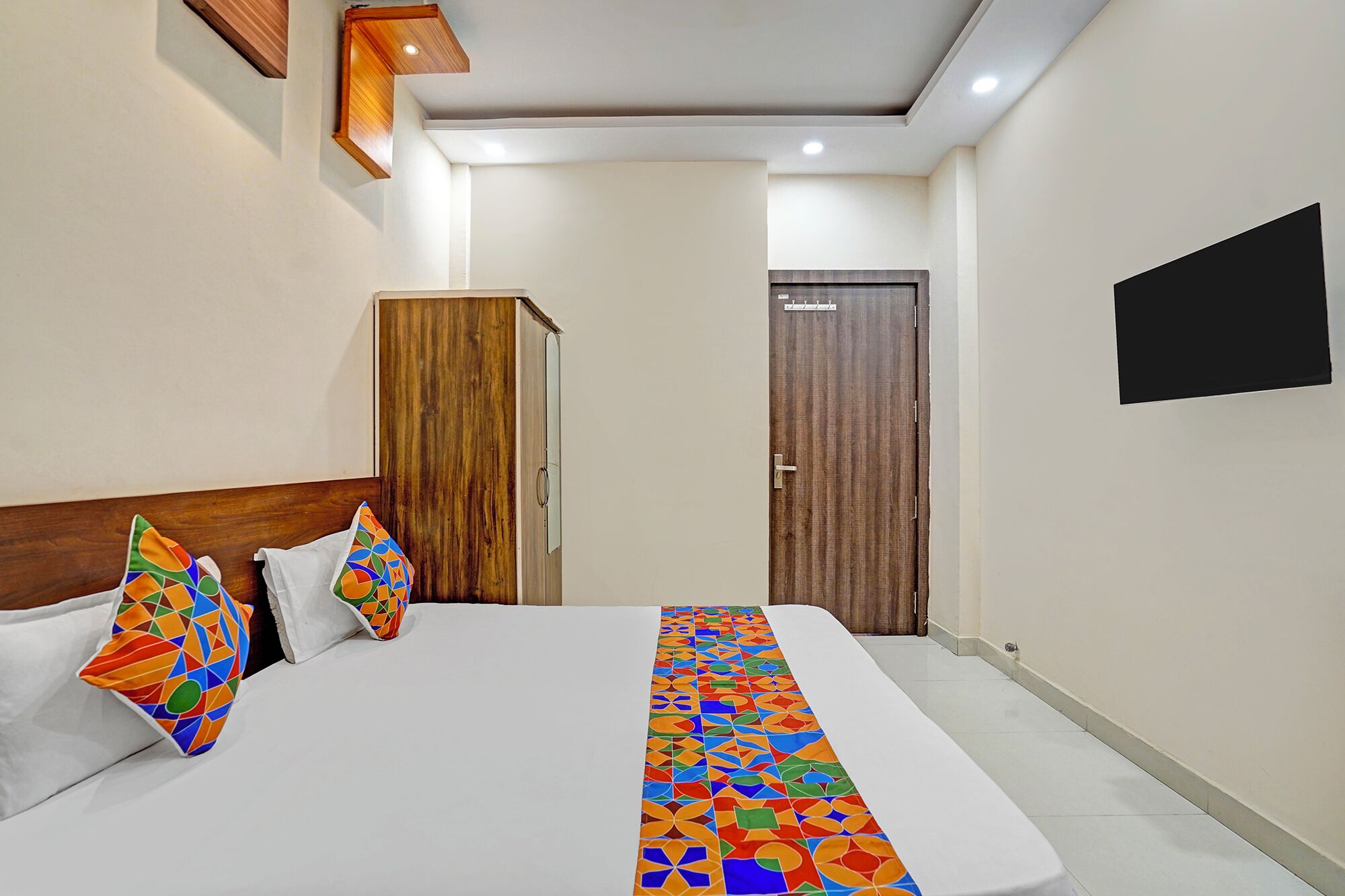 A hotel room with a bed, a wooden wardrobe, a door, and a TV mounted on the wall.