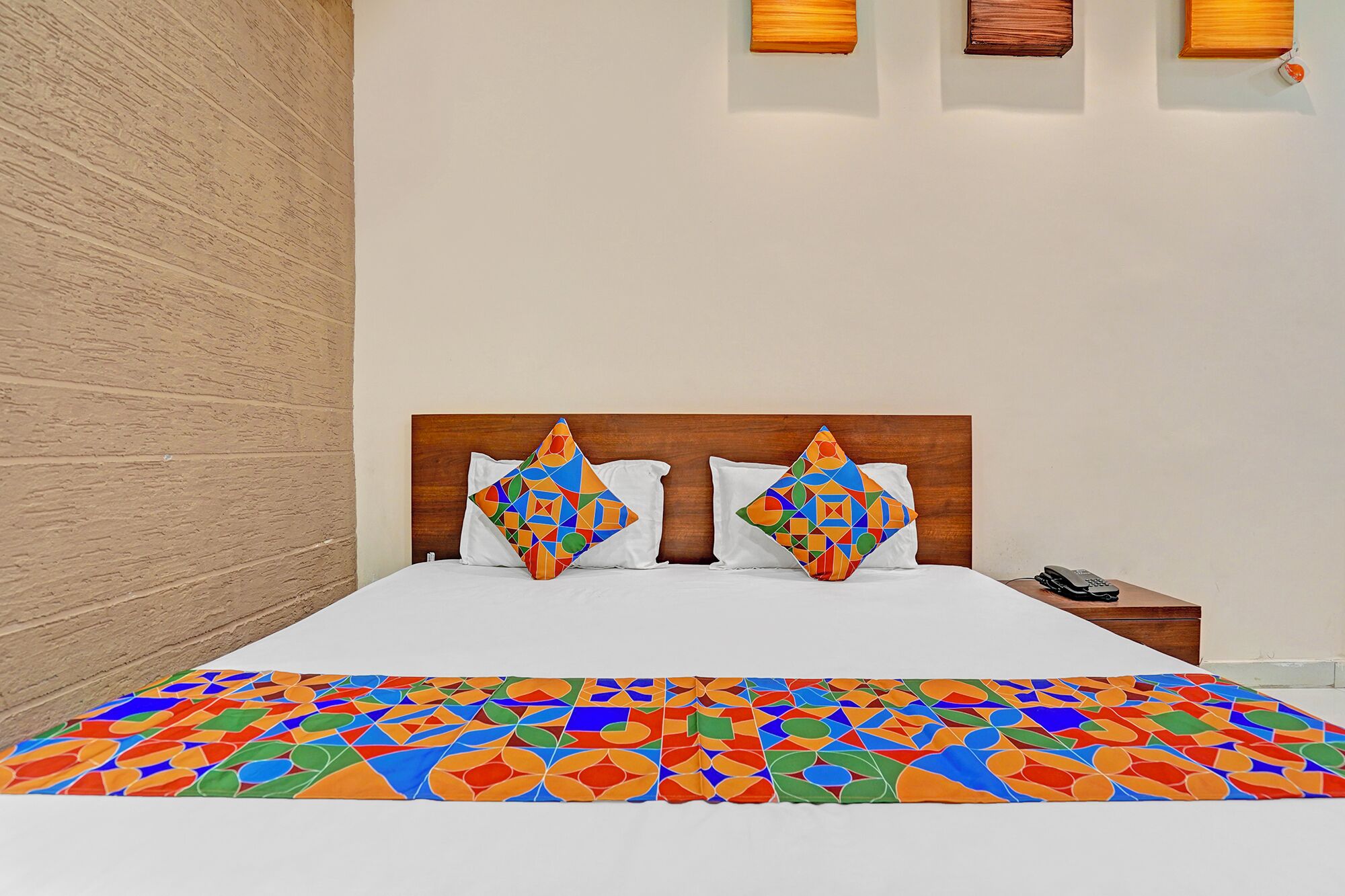 Habitación de hotel con cabecera de madera, dos almohadas de colores con motivos geométricos, una colcha blanca y una mesita de noche pequeña con un teléfono.