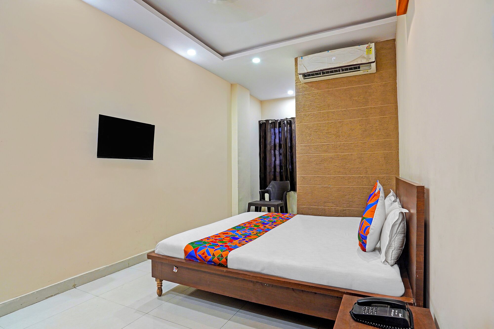 Une chambre d’hôtel comprenant un lit, une table de chevet avec un téléphone, un appareil de climatisation, une petite télévision et un rideau.
