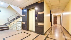 A long, well-lit hallway with a marble floor, a staircase on the left, and a door with an elevator button on the right. - Fabhotel The Grand Manmuralidhar (Jaipur)