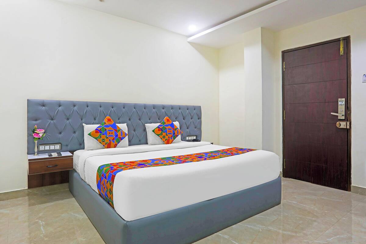 A bedroom with a tufted headboard, a bed with a colorful geometric-patterned bedspread, a bedside table with a vase of flowers, and a dark wooden door.