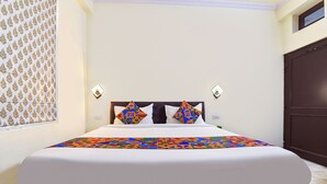A neatly made bed with a white bedspread and colorful geometric pillows, a dark wooden headboard, and a wall with patterned curtains. - Fabhotel Silver Stay 04 (Jaipur)