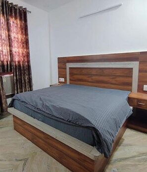 A bedroom with a wooden bed frame, a grey bedspread, a bedside table, and a wall-mounted light fixture. - Fabhotel Vibe Living (Gurugram)