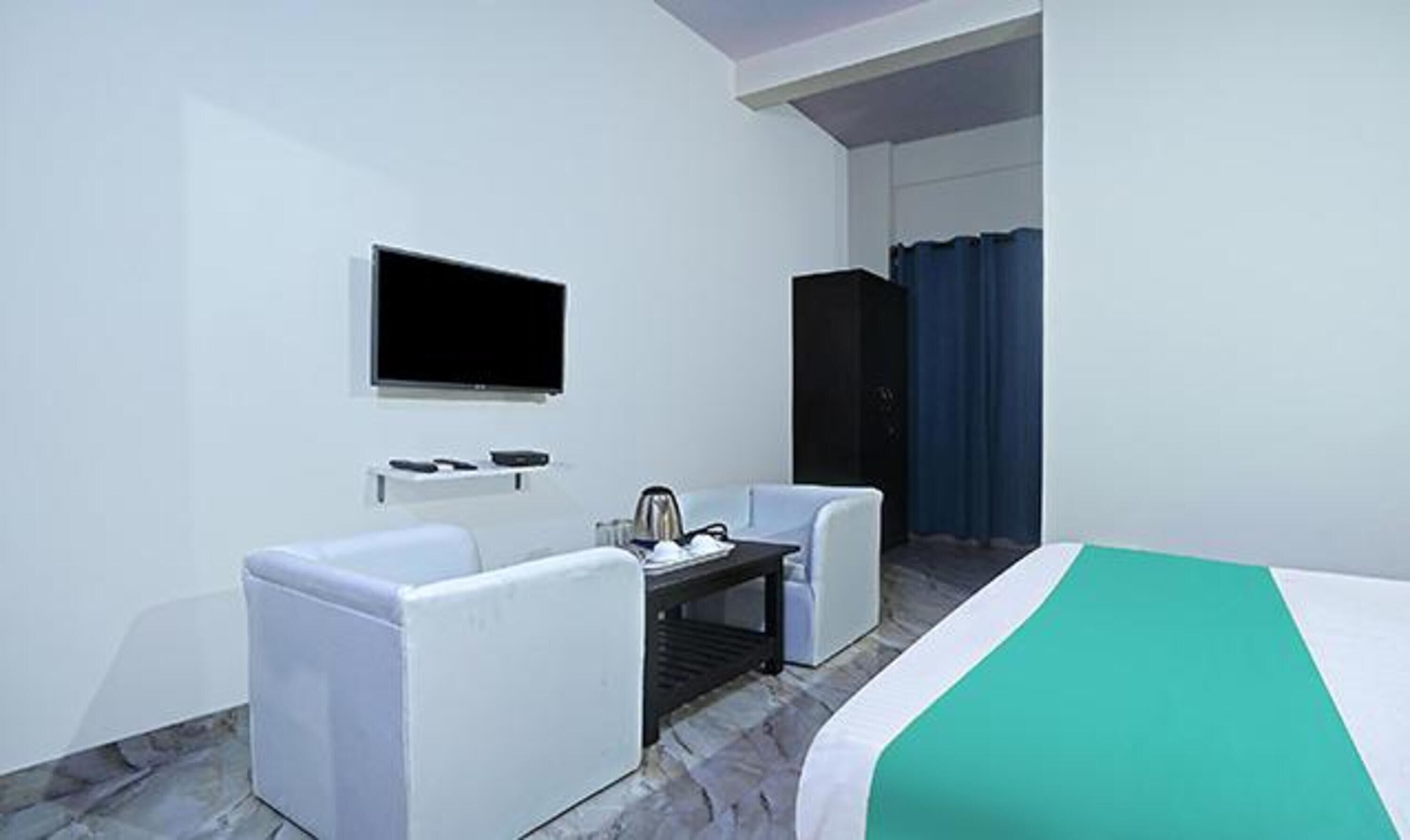 A hotel room with a bed, a TV mounted on the wall, a small table with a kettle, and a chair.