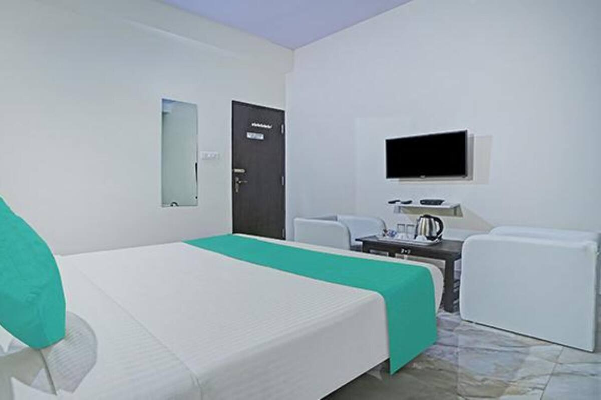 A hotel room with a bed, a desk with a chair, a TV mounted on the wall, and a door.