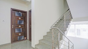 Una escalera con barandillas metálicas que sube a un piso superior, una puerta de madera con una placa de número y una pared de color claro.