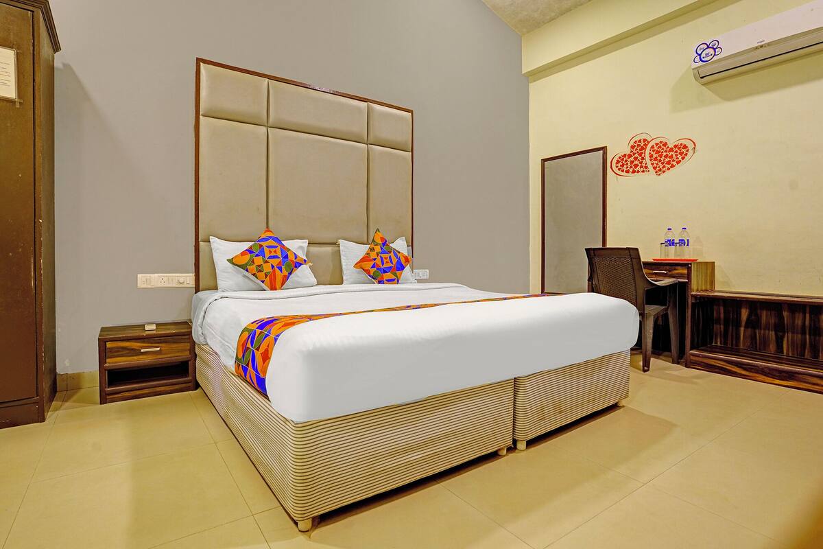 A hotel room with a large bed, a wooden nightstand, a desk with a chair, and a wall-mounted air conditioner.