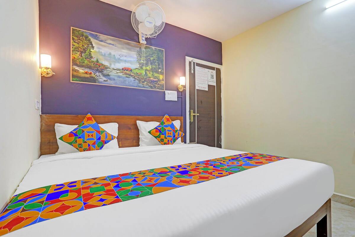 A hotel room with a bed, a painting on the wall, a fan, and a door with a sign.