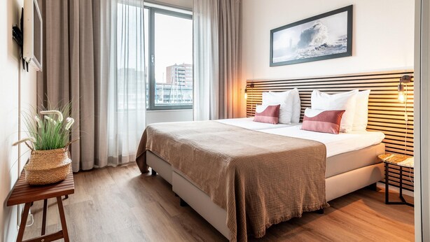 A modern hotel room with a large bed, a bedside table with a plant, a city view, and a framed picture on the wall. - Beautiful Apartment With Harbour Views (The Hague)