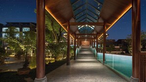 A covered walkway with a glass ceiling, leading to a pool area, surrounded by trees and buildings. - MOONRISE HOTELS & RESORTS (Deyang)