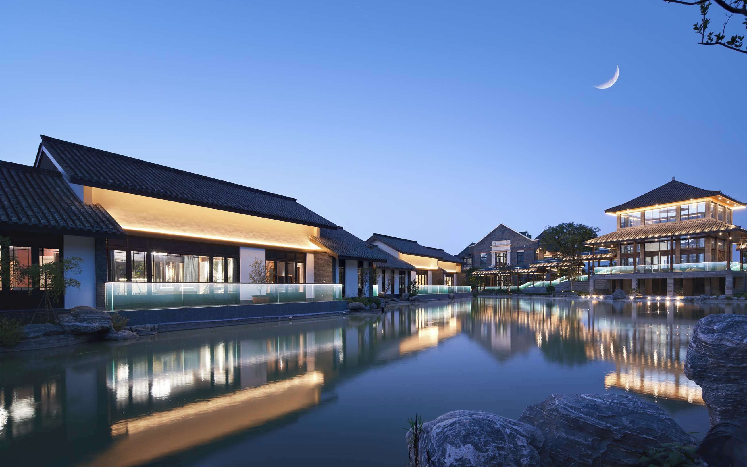 A serene hotel setting with traditional architecture, a reflective pool, and a crescent moon in the sky.