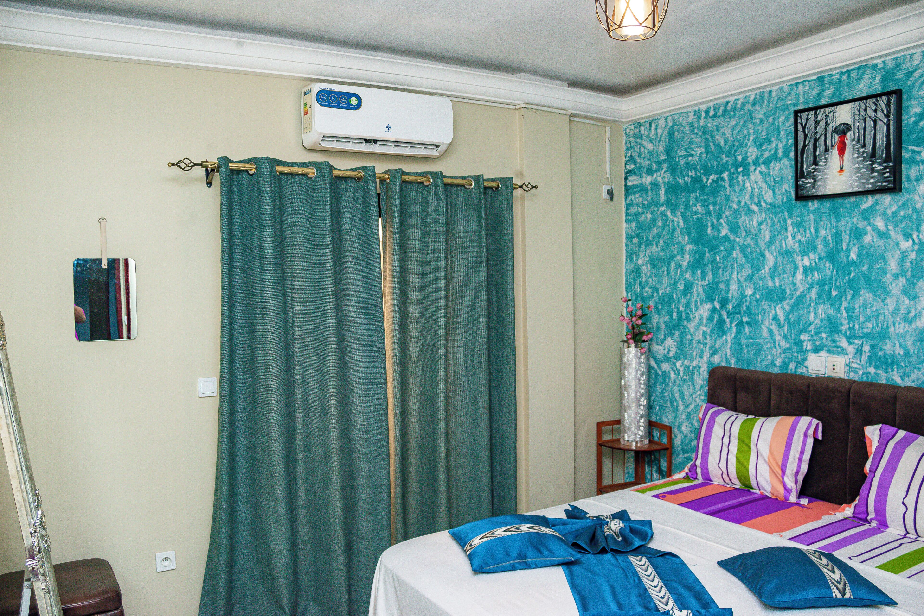 Una habitación de hotel con cama, cortinas, aire acondicionado, un cuadro enmarcado y un jarrón con flores.