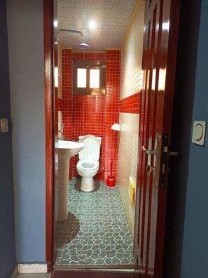 A bathroom with a white toilet, red tiled walls, a sink, and a shower. - Résidence JTC (Douala)